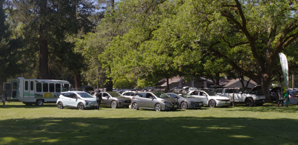 EV Showcase Ukiah Earth Day 2024 Many electric vehicles, including an electric bus, parked on the grass underneath a large oak tree.